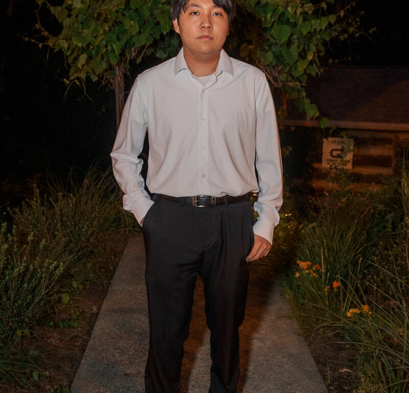 Young man in a white dress shirt and black pants stands on a garden path at night, hands in pockets, looking at the camera.