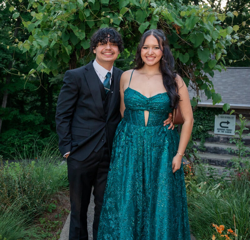 Two teenagers in formal attire posing outdoors; boy in black suit and teal tie, girl in teal sequined gown on a garden path.