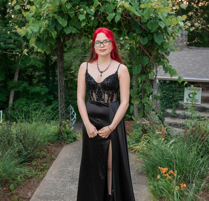 Young woman with bright red hair in a black lace bodice gown standing on a garden path under a leafy archway.
