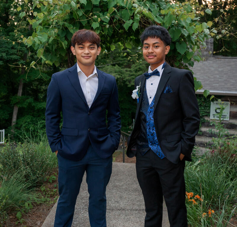 Two men in formal suits pose on a garden path, one in a navy suit and white shirt, the other in a black suit with a blue patterned vest.