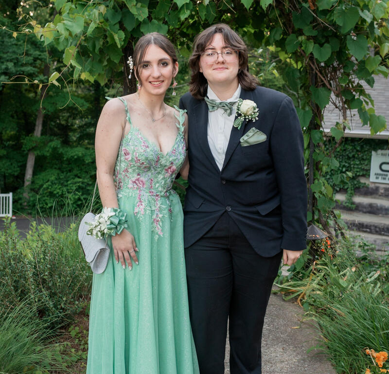 Two people in formal attire pose outdoors under a leafy arch: a woman in a mint green floral gown with a corsage and silver clutch, and a man in a black tuxedo with a green bow tie and boutonniere.