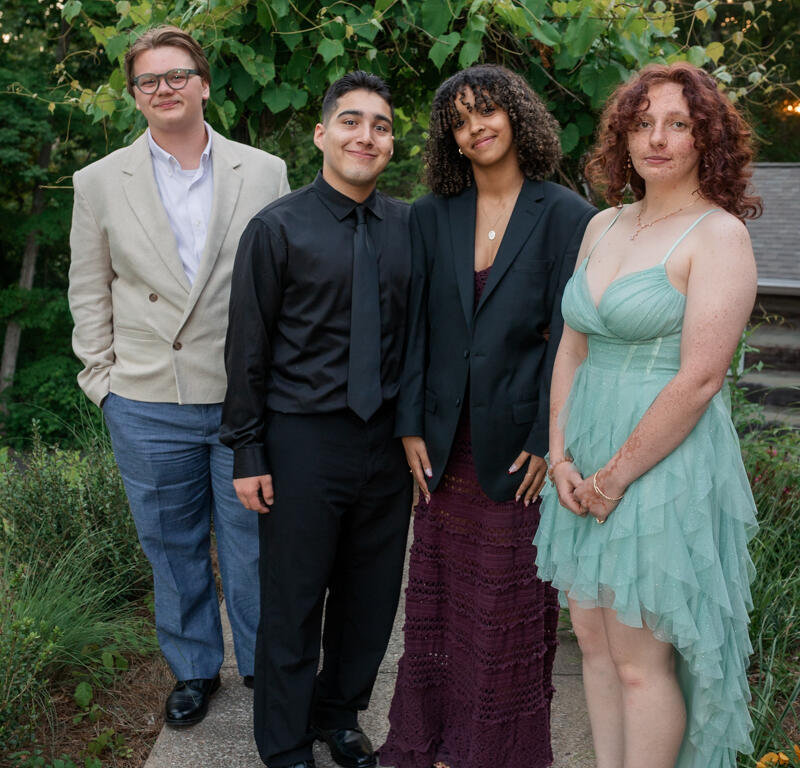 Four young people in formal attire pose on a garden path outdoors, smiling at the camera with greenery behind them.
