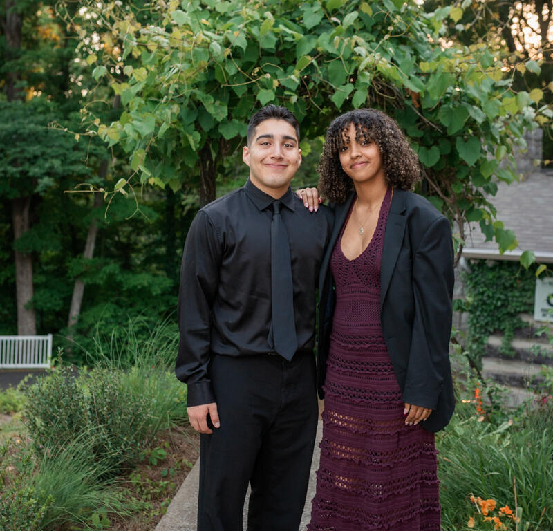 Couple posing for a portrait outdoors on a garden path, man in black shirt and tie, woman in a maroon lace dress and black blazer, smiles at camera.