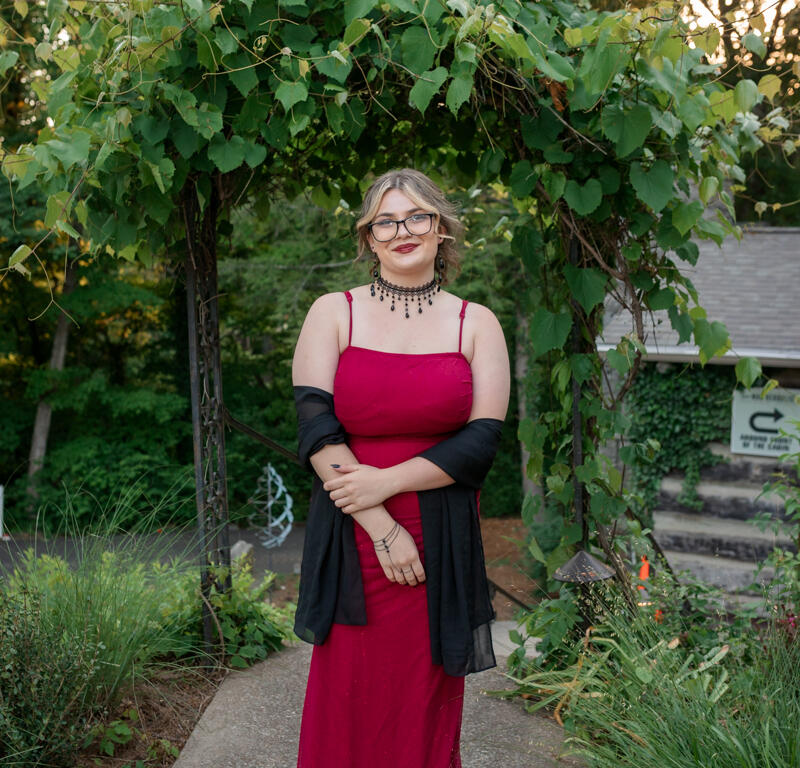 Woman in a magenta gown standing on a garden path beneath a leafy arch, wearing a black shawl and statement necklace.