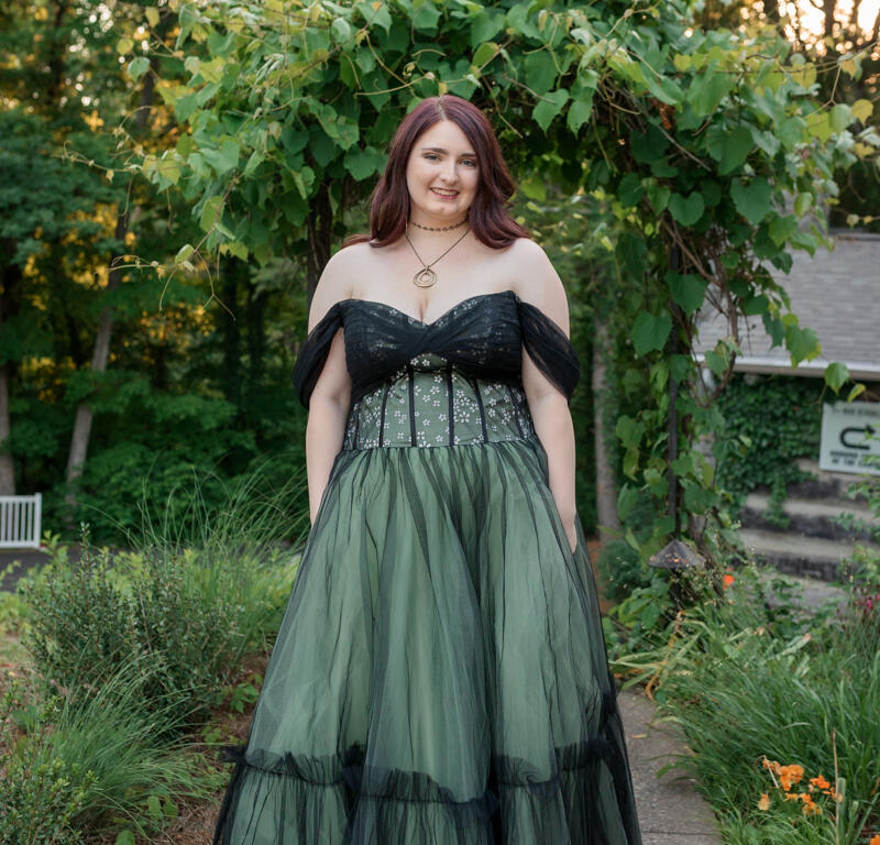 Woman in a strapless, dark bodice and layered emerald gown standing on a garden path, smiling at the camera.