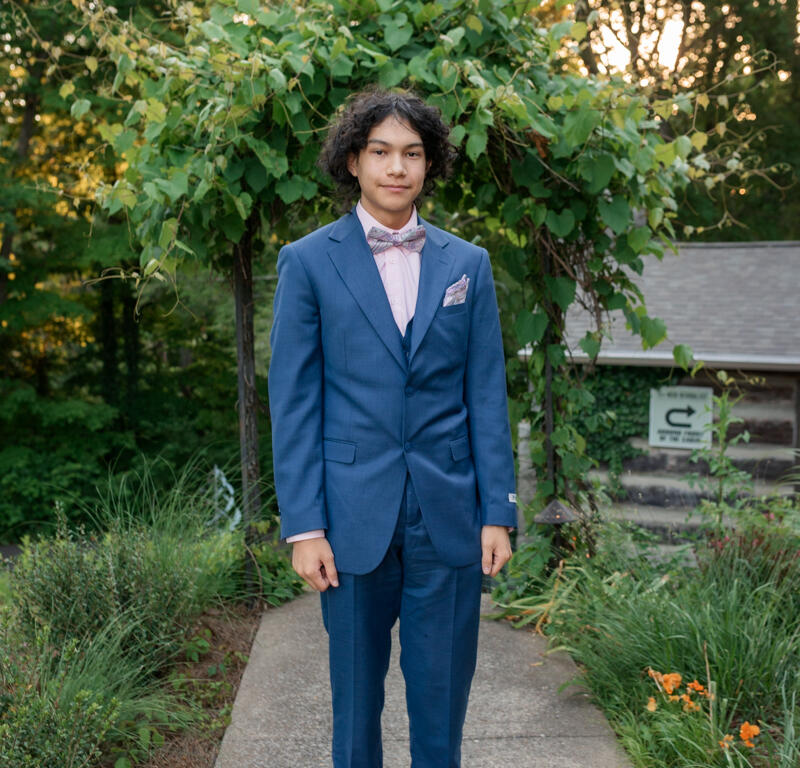 Young man in a blue suit with a pink shirt and bow tie standing on a garden path, greenery behind him.