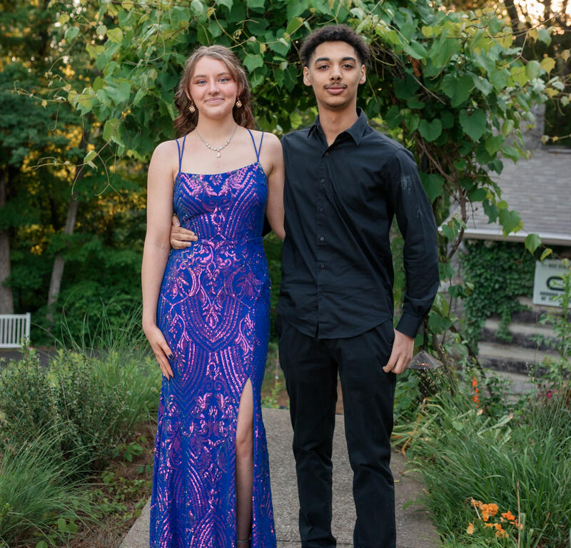 Young couple posing on a garden path: woman in a blue sequined gown with a thigh-high slit and a man in a black dress shirt and pants beside her.