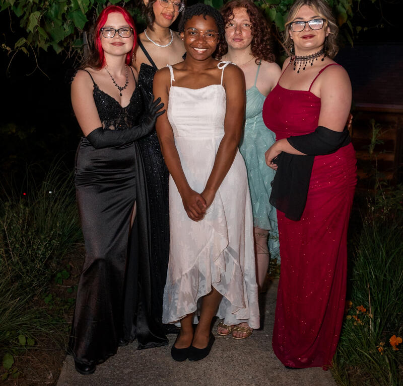 Group of five young women in formal dresses posing on a garden path at night, smiling under leafy backdrop.