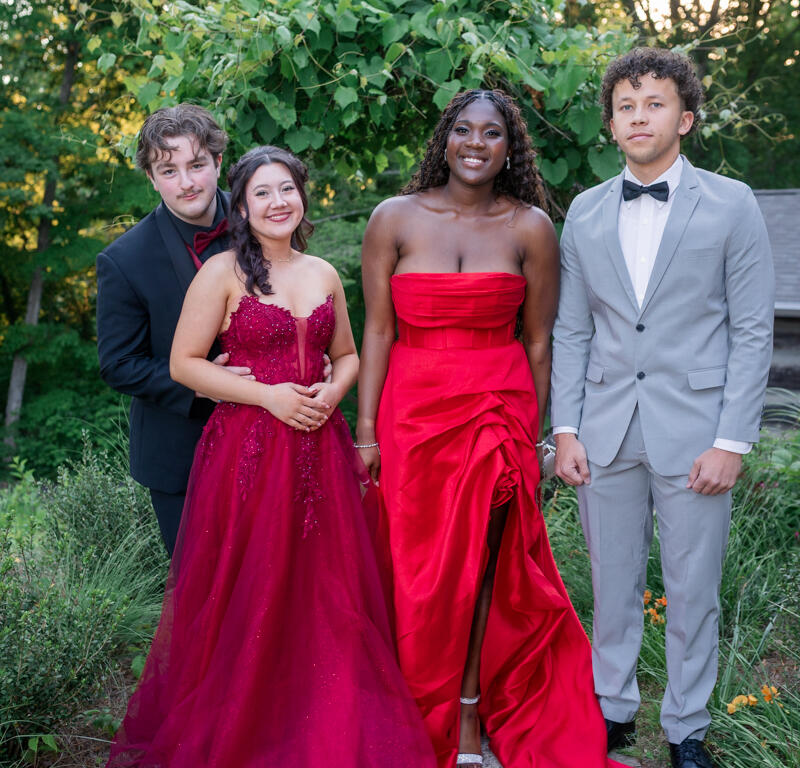 Four friends in formal attire pose together on a garden path, smiling at the camera.