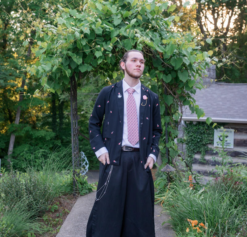 Man in a long black novelty coat and wide trousers stands on a garden path, posing beneath a leafy arch.