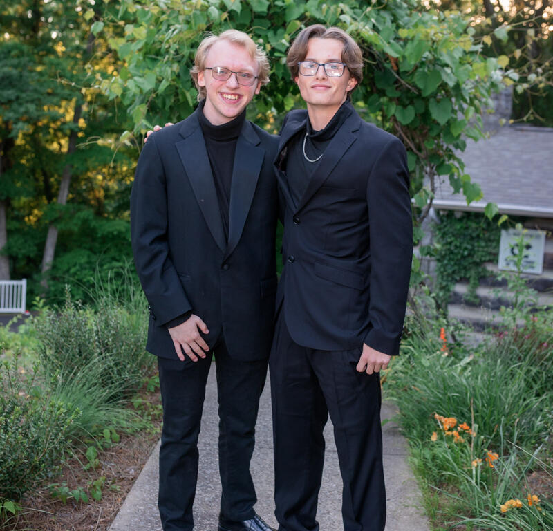 Two young men in black suits posing on a garden path, smiling with greenery behind them.