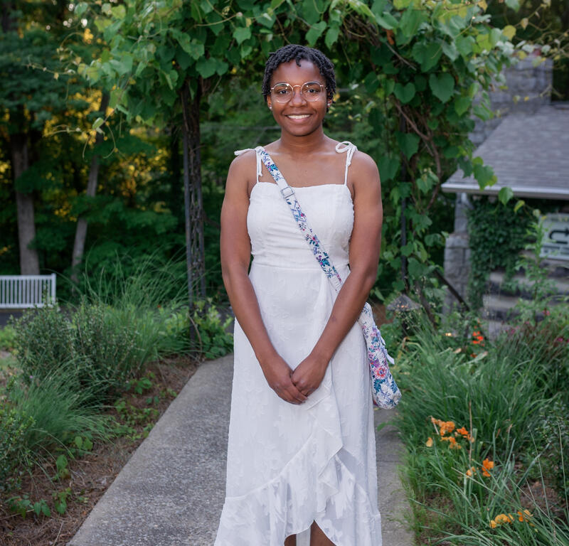 Smiling woman in a white summer dress with a floral crossbody bag standing on a garden path.