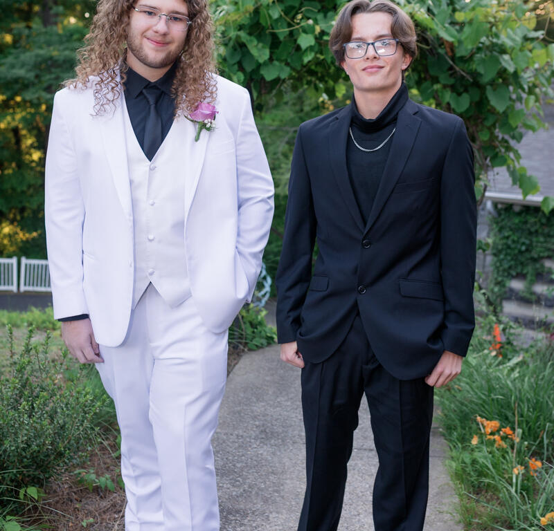 Two young men in formal suits pose on a garden path: one in a white suit with a pink boutonniere, the other in a black suit with a chain necklace and glasses.