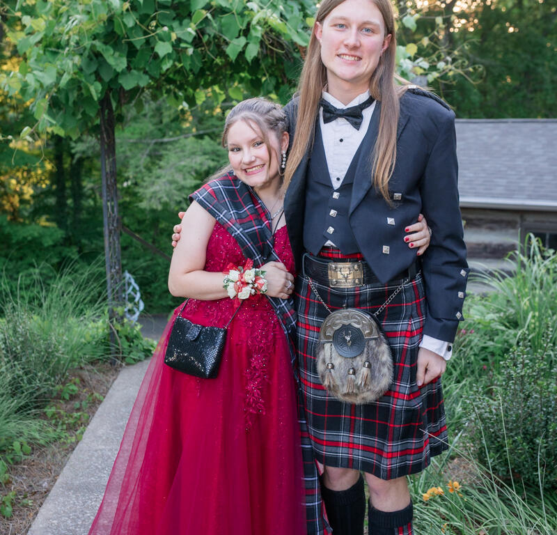 Two teens in formal wear pose together outdoors: a girl in a red ball gown with a corsage and small black purse and a boy in a black tuxedo with a red plaid kilt and a furry sporran, smiling under green trees.