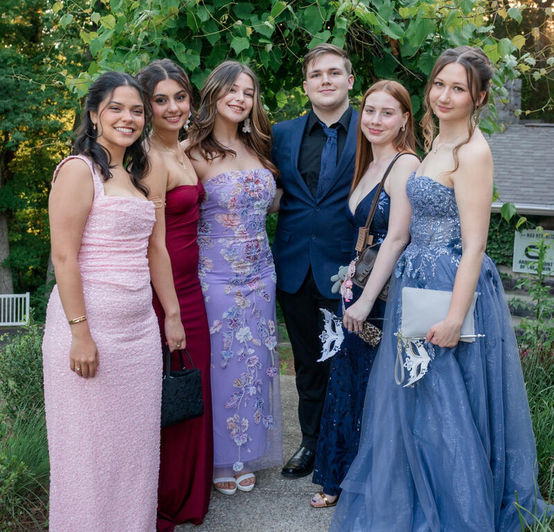 Group of six friends in formal attire posing outdoors for a prom photo on a garden path.