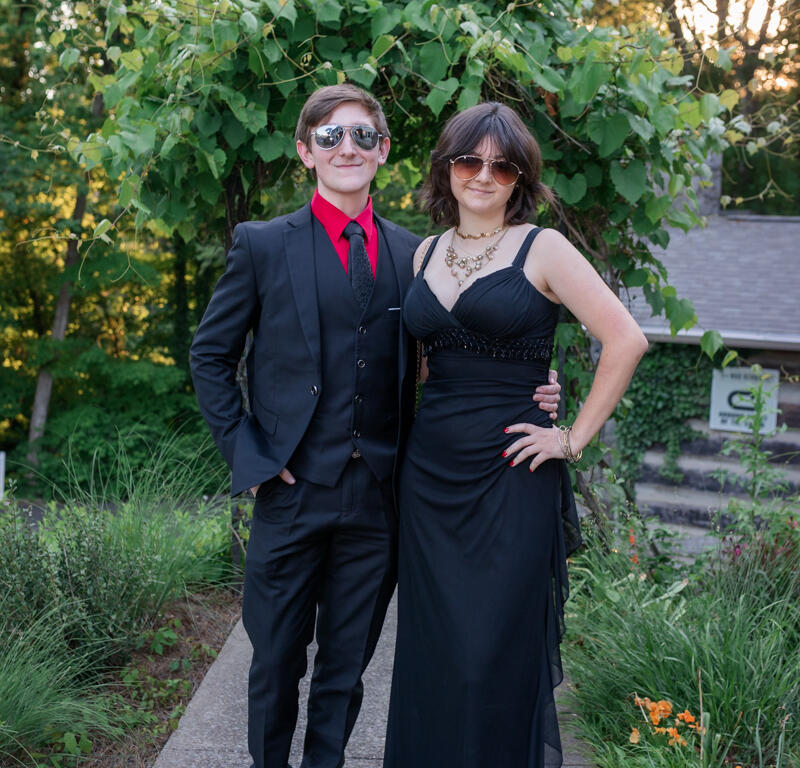 Couple in formal attire posing outdoors in a garden, man in a black suit with red shirt and tie, woman in a black gown with sunglasses and jewelry.