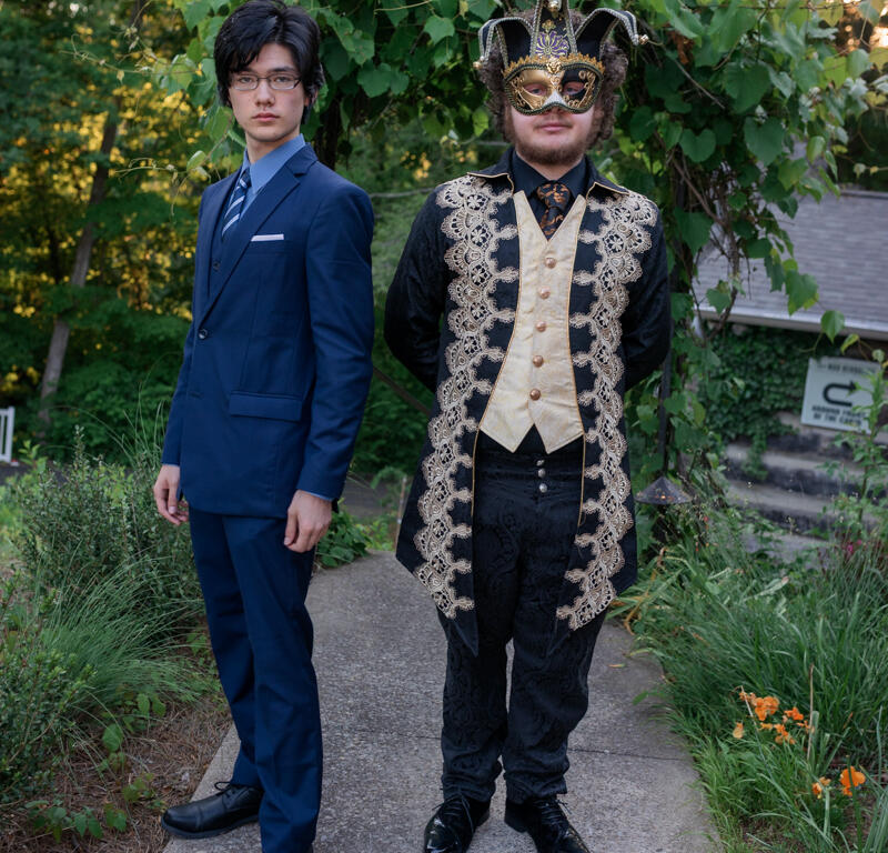 Two men stand on a garden path: one in a blue suit with glasses, the other in a gold-trimmed masquerade costume and mask.
