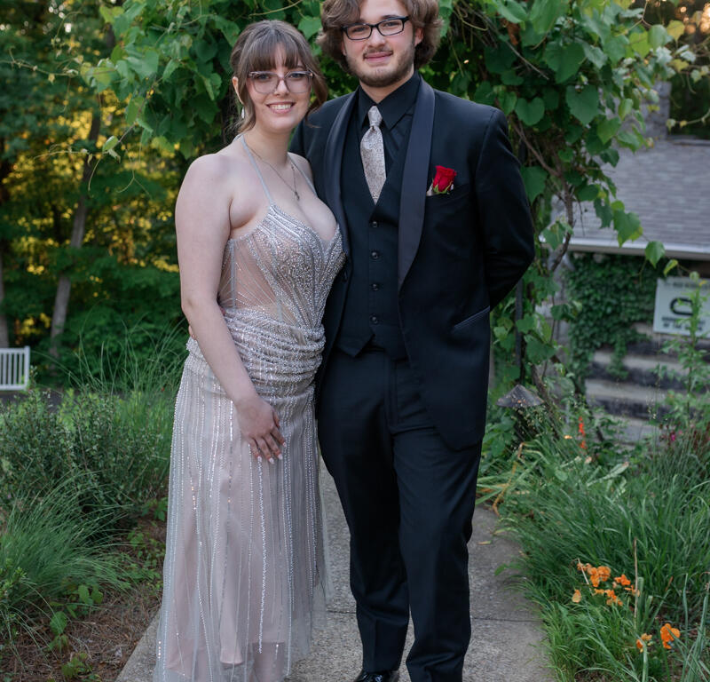 Couple dressed in formal attire posing on a garden path with greenery in the background, the woman in a beaded gown and the man in a black suit with a red boutonniere.