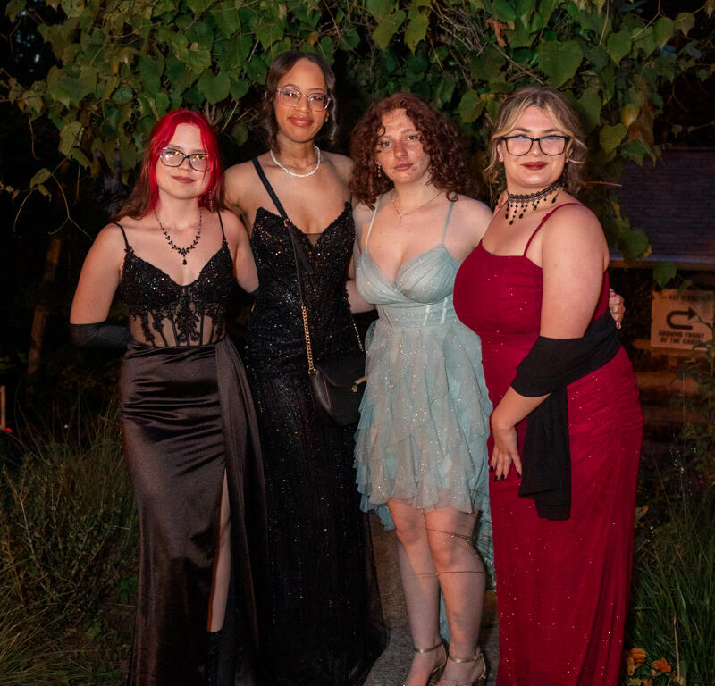 Four young women in formal gowns pose together outdoors at night, smiling under a leafy backdrop.