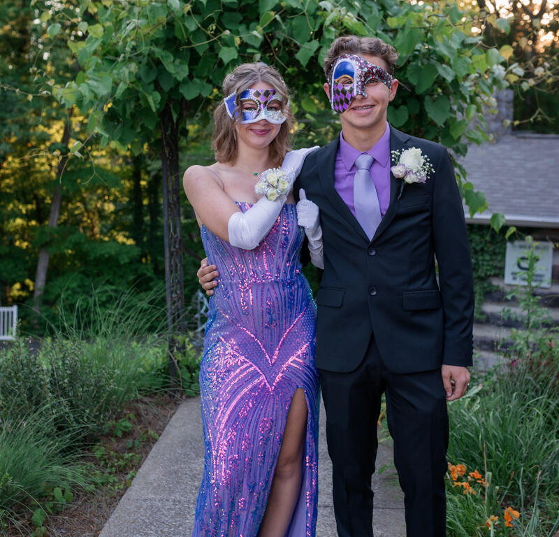 Couple in masquerade masks posing outdoors; woman in a purple sequin gown with a slit and a man in a dark suit with a purple tie.