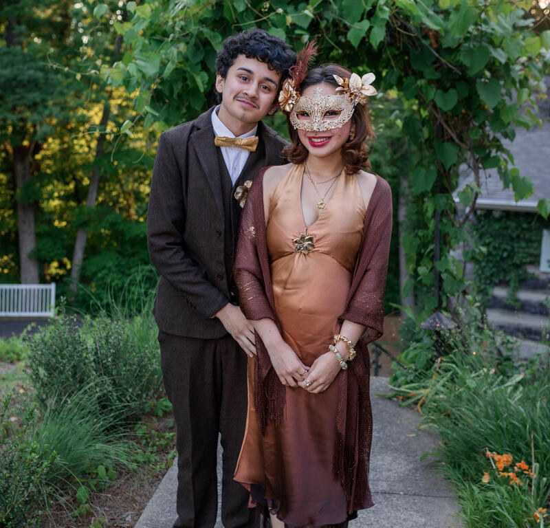 Couple in vintage attire posing together outdoors under a green leafy arch; man in brown suit with a gold bow tie, woman in bronze dress and ornate mask.
