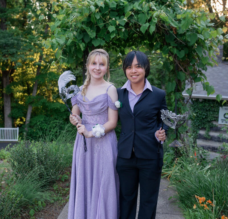 Two teens in formal dress pose outdoors under a leafy garden arch, each holding a silver masquerade mask.