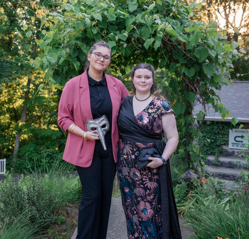 Two people pose on a garden path beneath a leafy arch; the left person wears a pink blazer and black outfit, holding a decorative mask, while the right person wears a black floral gown.