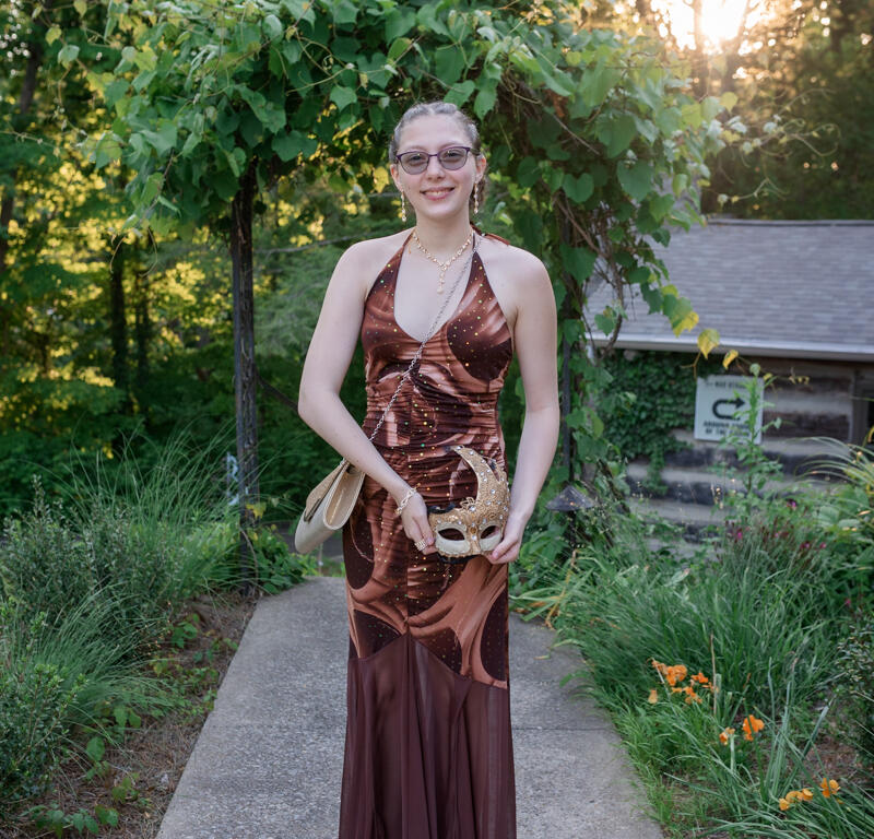 Person in a bronze-brown halter gown standing on a garden path, holding a decorative masquerade mask.
