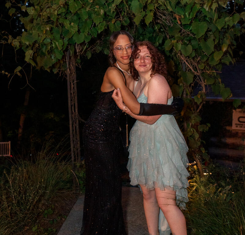 Two women in formal gowns embrace on a garden path at night under leafy overhead branches.