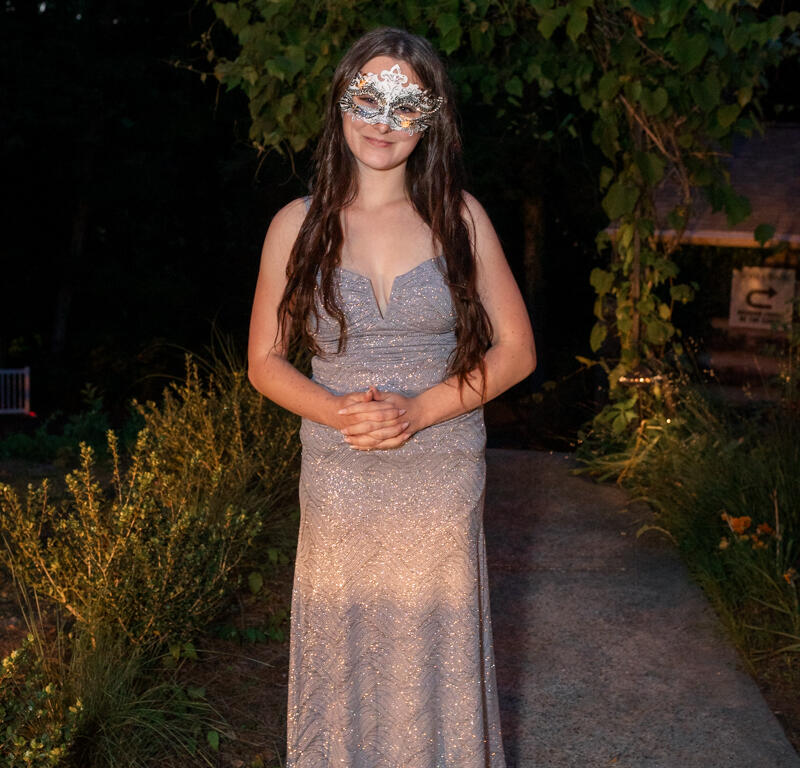 Woman in a glittery silver evening gown and a decorative masquerade mask stands on a stone garden path at night.