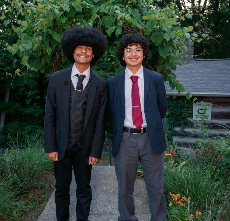 Two young men in suits pose on a garden path, smiling, with leafy shrubs behind them.
