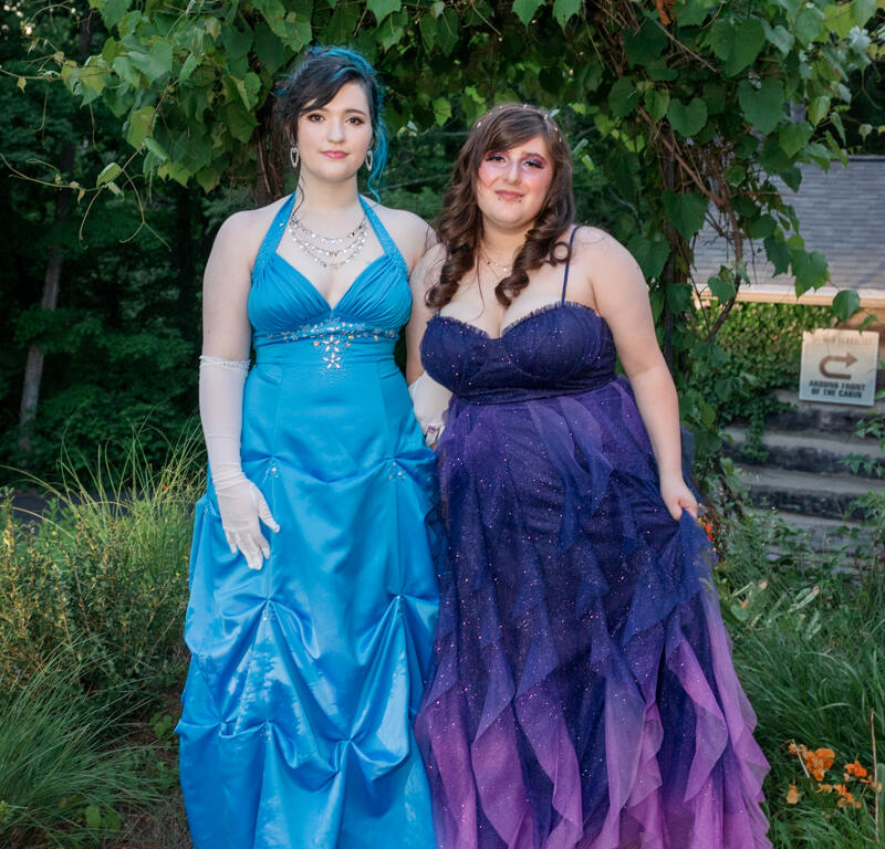 Two young women in formal gowns posing on a garden path under a leafy archway.