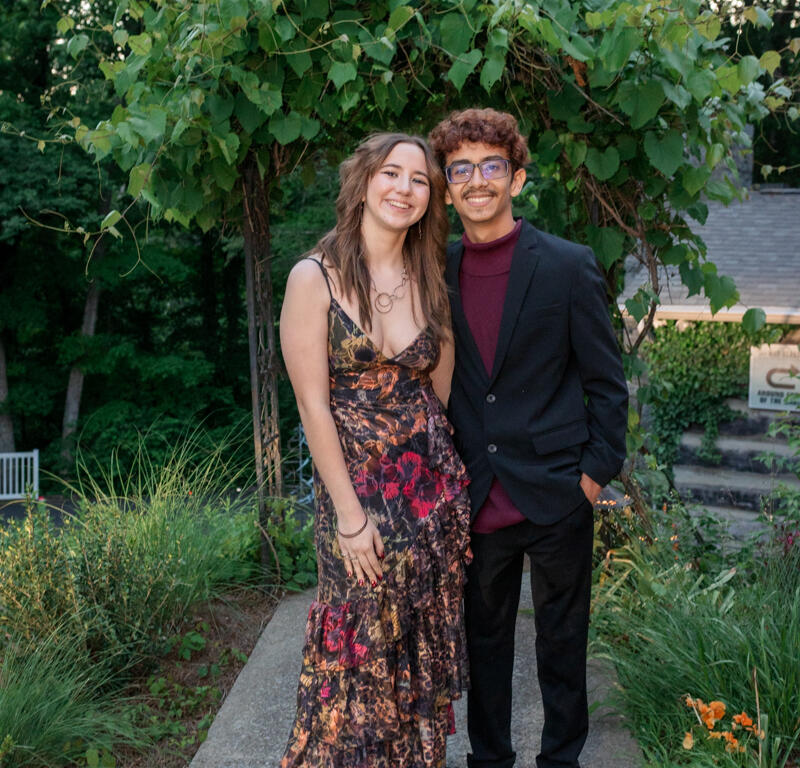 Smiling young couple posing on a garden path, the woman in a floral, layered dress and the man in a dark blazer with a maroon sweater beneath it.