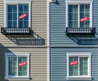 Four exterior windows on a beige and blue duplex with red SOLD signs in the panes, indicating sold properties in each unit.