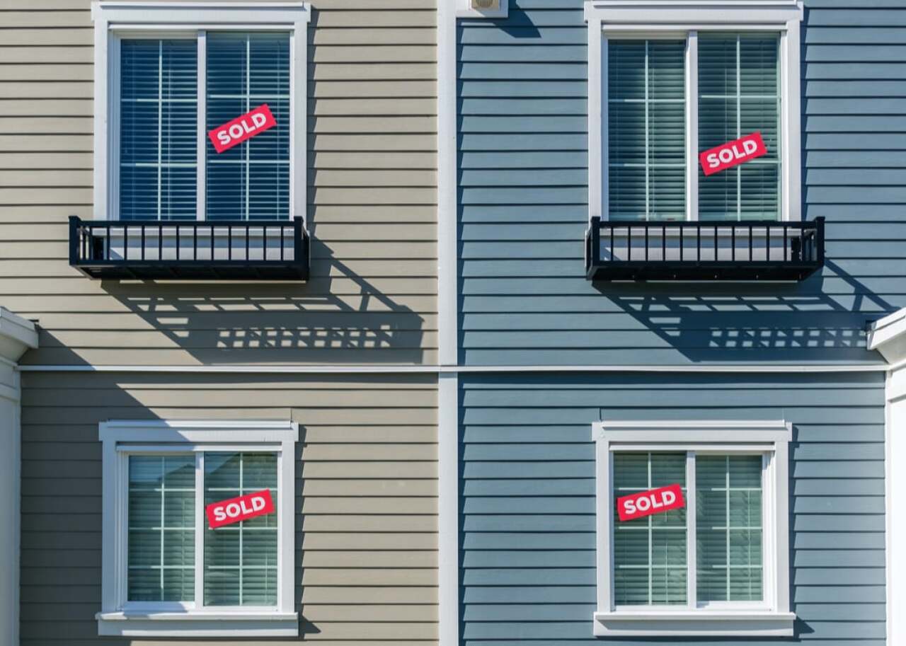 Four exterior windows on a beige and blue duplex with red SOLD signs in the panes, indicating sold properties in each unit.