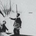 Black-and-white winter scene: a woman in a long skirt stands on a curb while three children in coats stand nearby, one child pointing toward her.