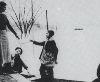 Black-and-white winter scene: a woman in a long skirt stands on a curb while three children in coats stand nearby, one child pointing toward her.
