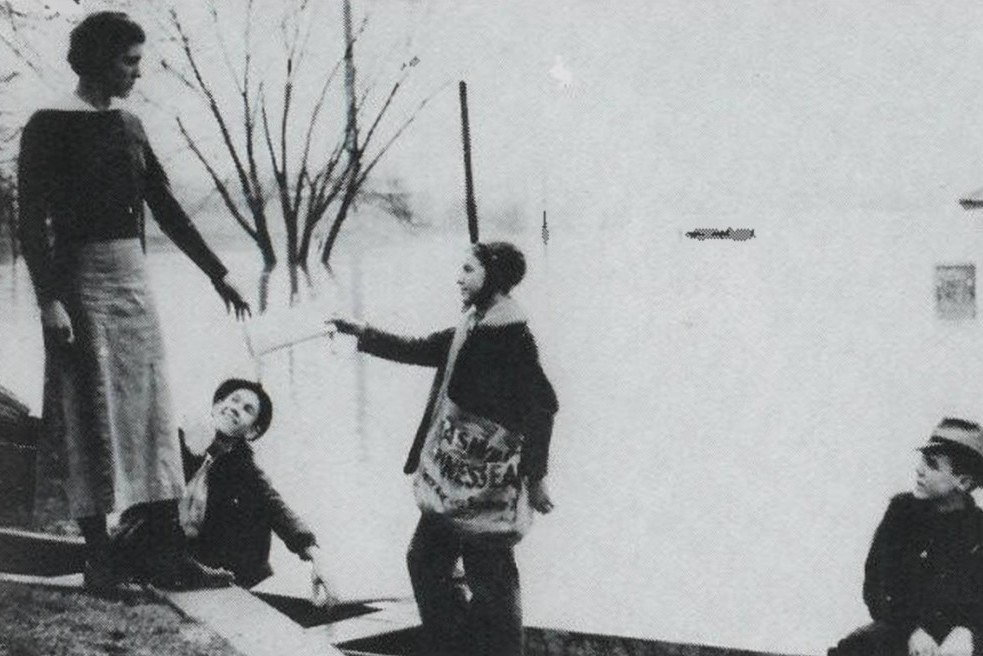 Black-and-white winter scene: a woman in a long skirt stands on a curb while three children in coats stand nearby, one child pointing toward her.