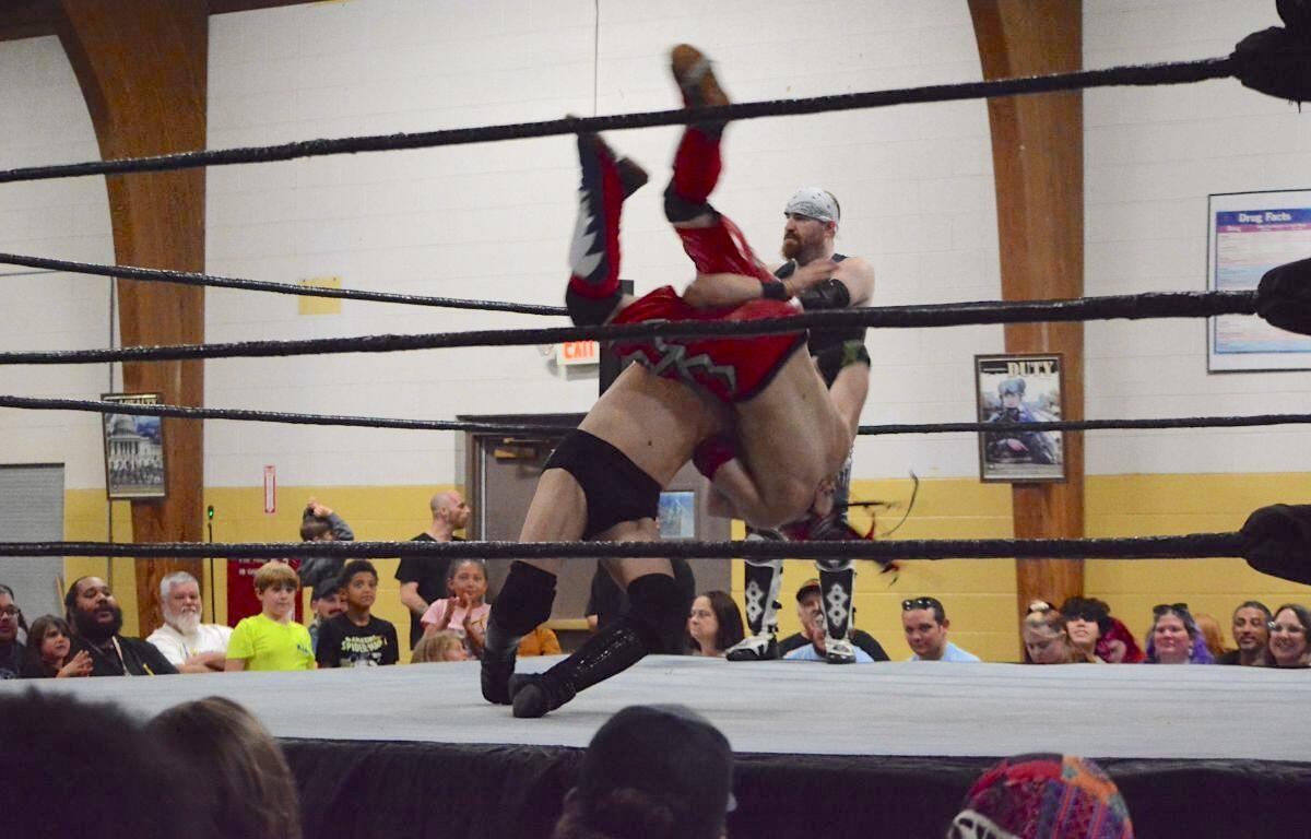 Two wrestlers clash in a ring as one lifts the other overhead during a match, with a crowd watching closely nearby.