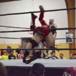 Two wrestlers clash in a ring as one lifts the other overhead during a match, with a crowd watching closely nearby.