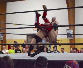 Two wrestlers clash in a ring as one lifts the other overhead during a match, with a crowd watching closely nearby.