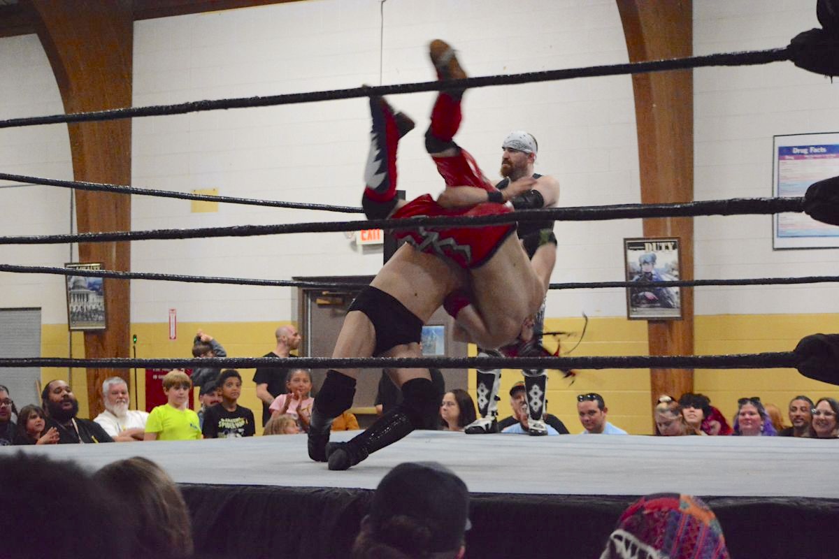 Two wrestlers clash in a ring as one lifts the other overhead during a match, with a crowd watching closely nearby.