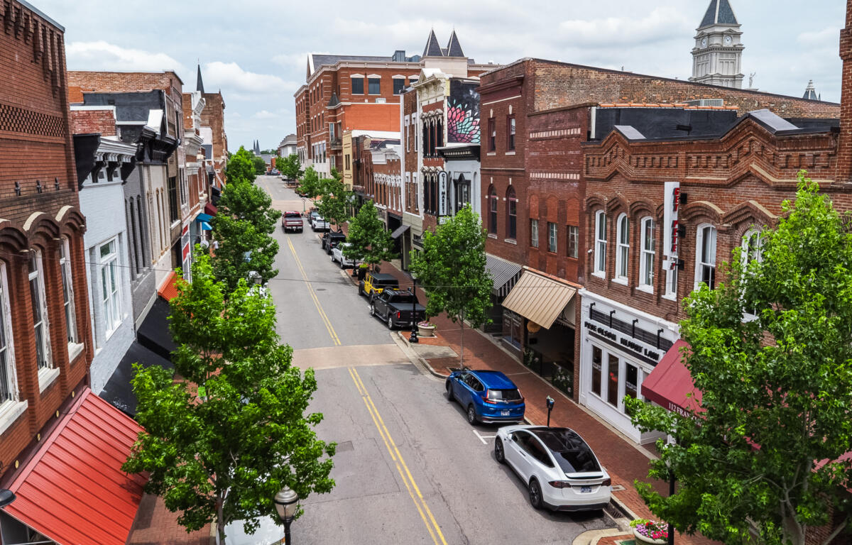 Bird's-eye view of a brick storefront district: a tree-lined street with parked cars and awning-covered shops on both sides.