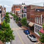 Bird's-eye view of a brick storefront district: a tree-lined street with parked cars and awning-covered shops on both sides.