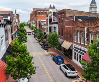 Bird's-eye view of a brick storefront district: a tree-lined street with parked cars and awning-covered shops on both sides.