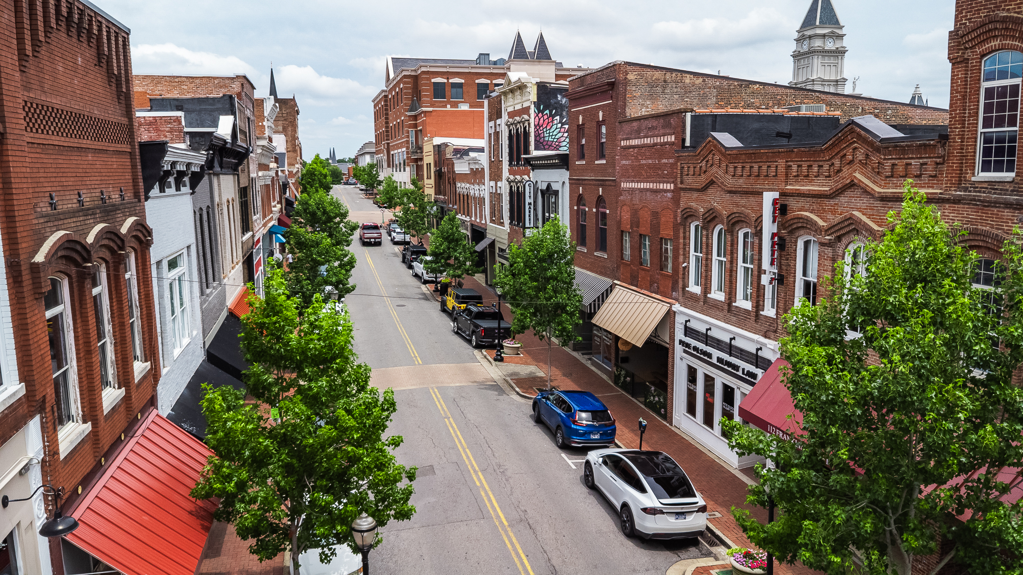 Bird's-eye view of a brick storefront district: a tree-lined street with parked cars and awning-covered shops on both sides.