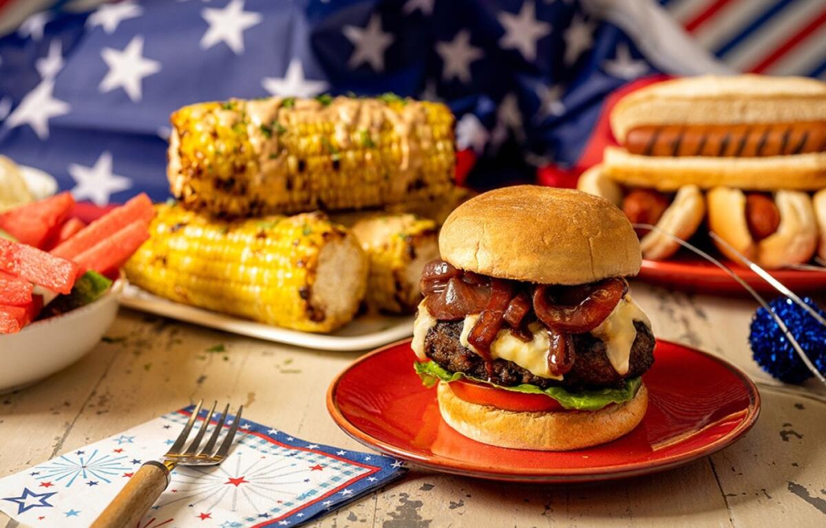 Patriotic barbecue spread featuring a cheeseburger, corn on the cob, watermelon, and hot dogs against an American flag backdrop.