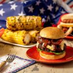 Patriotic barbecue spread featuring a cheeseburger, corn on the cob, watermelon, and hot dogs against an American flag backdrop.