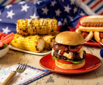 Patriotic barbecue spread featuring a cheeseburger, corn on the cob, watermelon, and hot dogs against an American flag backdrop.