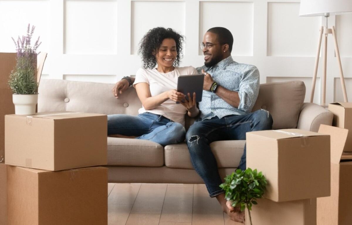 Couple sits on a beige couch amid unpacked boxes, smiling at a tablet together in a new home.